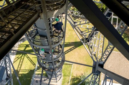 Menschen mit grünen Helmen steigen eine Wendeltreppe der Eisenbahnhochbrücke Rendsburg hinauf. Die Metallstruktur wirft Schatten auf den Boden.