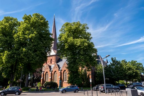 Die Sankt Martin Kirche in Nortorf, umgeben von grünen Bäumen und parkenden Autos, unter einem klaren blauen Himmel.