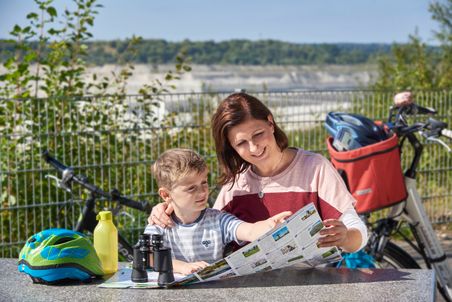Eine Frau und ein Junge studieren eine Karte an einem Tisch im Freien. Neben ihnen liegen ein Fahrradhelm, eine Trinkflasche und ein Fernglas.