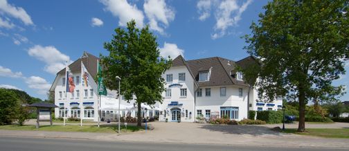 Das Hotel Wikingerhof zeigt sich mit weißer Fassade und mehreren Fahnen im Vordergrund. Ein Baum steht vor dem Gebäude, der Himmel ist blau mit wenigen Wolken.