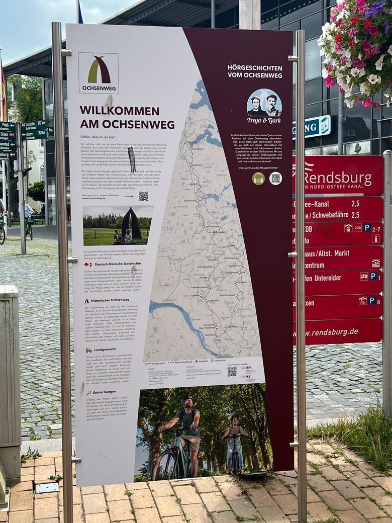 Ein Informationsschild in Rendsburg begrüßt Besucher am Ochsenweg. Es zeigt eine Karte und bietet Informationen zur Geschichte und Sehenswürdigkeiten der Region.