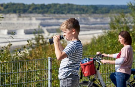 Ein Junge beobachtet mit einem Fernglas die Umgebung, während eine Frau mit Fahrrad im Hintergrund steht. Im Hintergrund ist die Kreidegrube Lägerdorf und Wald zu sehen.