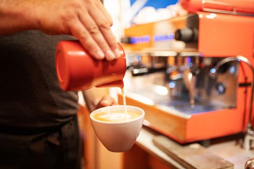 Ein Barista gießt Milch in eine Tasse Kaffee, während im Hintergrund eine orange Espressomaschine zu sehen ist.