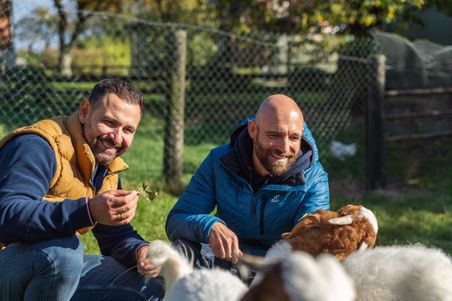 Zwei Männer füttern auf dem Eichenhof Grauel am Ochsenweg Ziegen im Garten und lächeln in die Kamera.