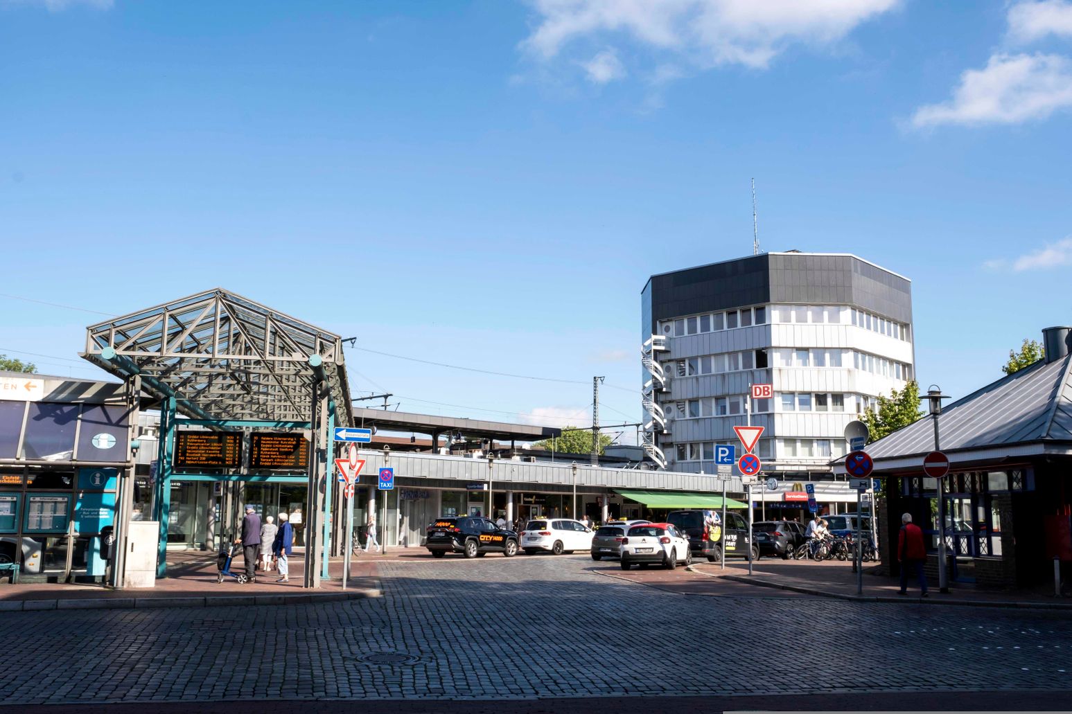 Der Bahnhof Neumünster mit einem modernen Glasdach und umliegenden Gebäuden. Autos und Menschen beleben die Szene bei sonnigem Wetter.