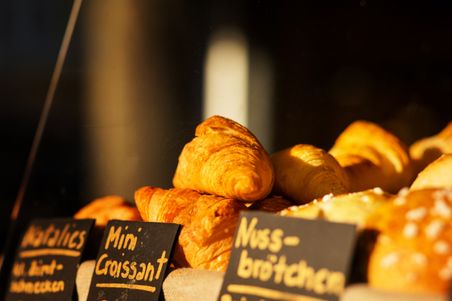 Frische Croissants und Brötchen in einer Bäckerei-Auslage, beschriftet mit handgeschriebenen Schildern. Sonnenlicht beleuchtet die goldene Kruste.