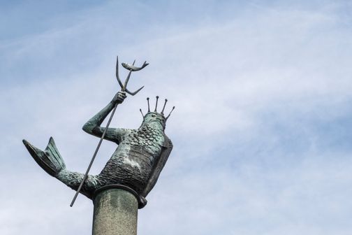 Die Statue von Neptun in Rendsburg zeigt den Meeresgott mit Dreizack und Krone, auf einer Säule sitzend, vor einem klaren blauen Himmel.