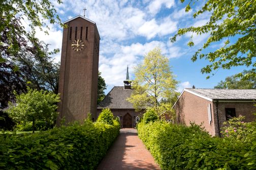 Die Kirche in Schacht-Audorf mit ihrem markanten Turm und einer Uhr, umgeben von grünen Bäumen und einem gepflegten Gartenweg.