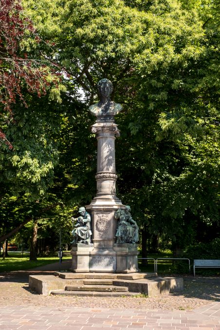 Ein Denkmal im Stadtpark Rendsburg zeigt eine Büste auf einer Säule, flankiert von zwei sitzenden Figuren. Umgeben von üppigem Grün.