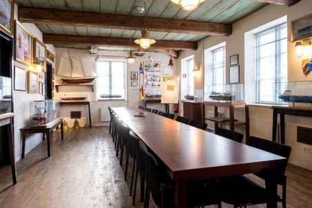 Ein Raum im Schifffahrtsarchiv Rendsburg mit einem langen Tisch, umgeben von Stühlen. An den Wänden sind Schiffsmodelle und Bilder ausgestellt.