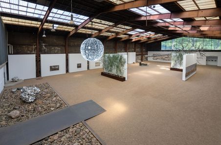 Große Ausstellungshalle mit moderner Kunstinstallation. Eine leuchtende Kugel hängt von der Decke, umgeben von Skulpturen und Pflanzenwänden.