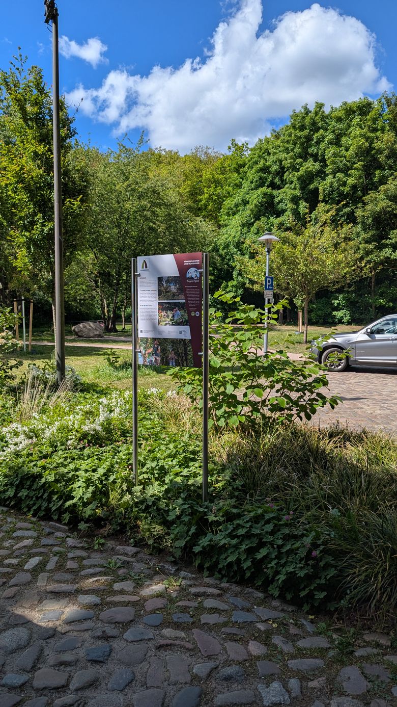 Ein Informationsschild zum Ochsenweg steht in einem grünen Park. Im Hintergrund ist ein Auto zu sehen. Der Himmel ist blau mit einigen Wolken.
