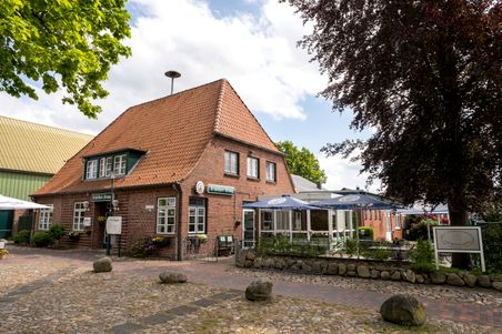 Das Bild zeigt die Gaststätte Schülper Kroog, ein charmantes Backsteingebäude mit rotem Ziegeldach und Außenterrasse, umgeben von Bäumen.