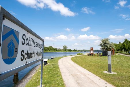 Schild am Schiffsanleger Schacht-Audorf, mit Blick auf einen Fluss und einen Weg, umgeben von grüner Landschaft und blauem Himmel.