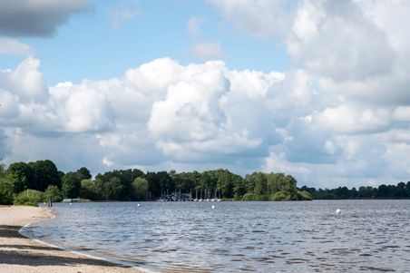 Ein idyllischer See mit Sandstrand und Segelbooten am Ufer. Im Hintergrund sind Bäume und ein bewölkter Himmel zu sehen.