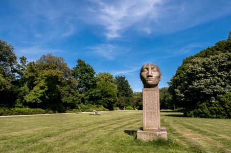 Eine große Steinskulptur eines Kopfes steht auf einem Sockel im Nortorfer Park. Umgeben von grünen Bäumen und Rasen, unter einem klaren blauen Himmel.