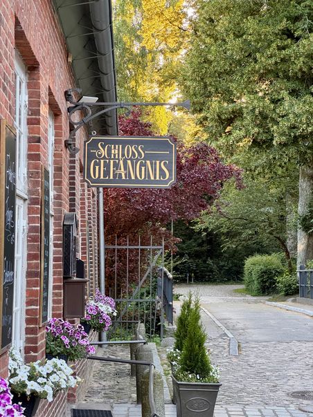 Eingang zum Schlossgefängnis mit einem dekorativen Schild und blühenden Blumen. Der Weg führt durch eine grüne, bewaldete Umgebung.