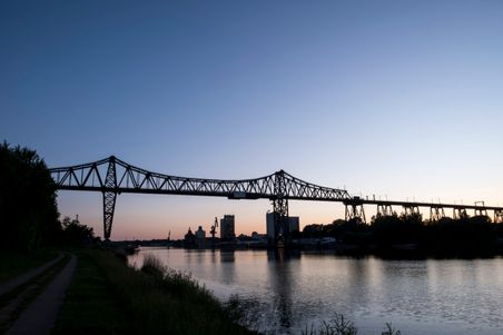 Die Eisenbahnhochbrücke Rendsburg erstreckt sich majestätisch über einen Fluss bei Sonnenuntergang, umgeben von einer ruhigen Landschaft.