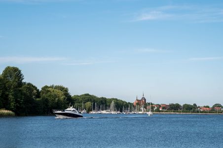 Ein Boot fährt durch den Obereiderhafen, im Hintergrund sind Segelboote und eine Kirche zu sehen. Der Himmel ist klar und blau.