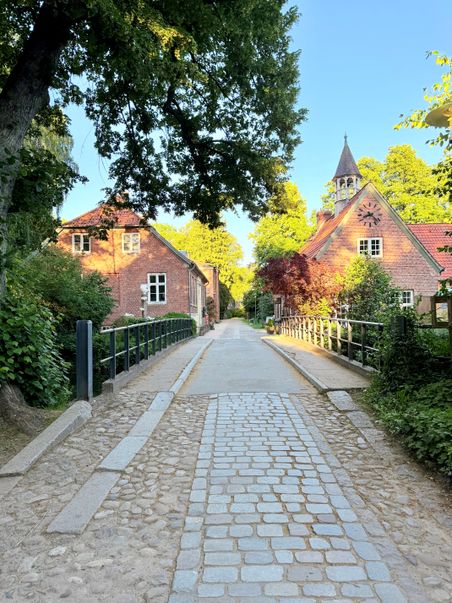Eine gepflasterte Brücke führt zu zwei roten Backsteingebäuden, umgeben von üppigem Grün. Ein Turm mit Uhr ragt im Hintergrund empor.
