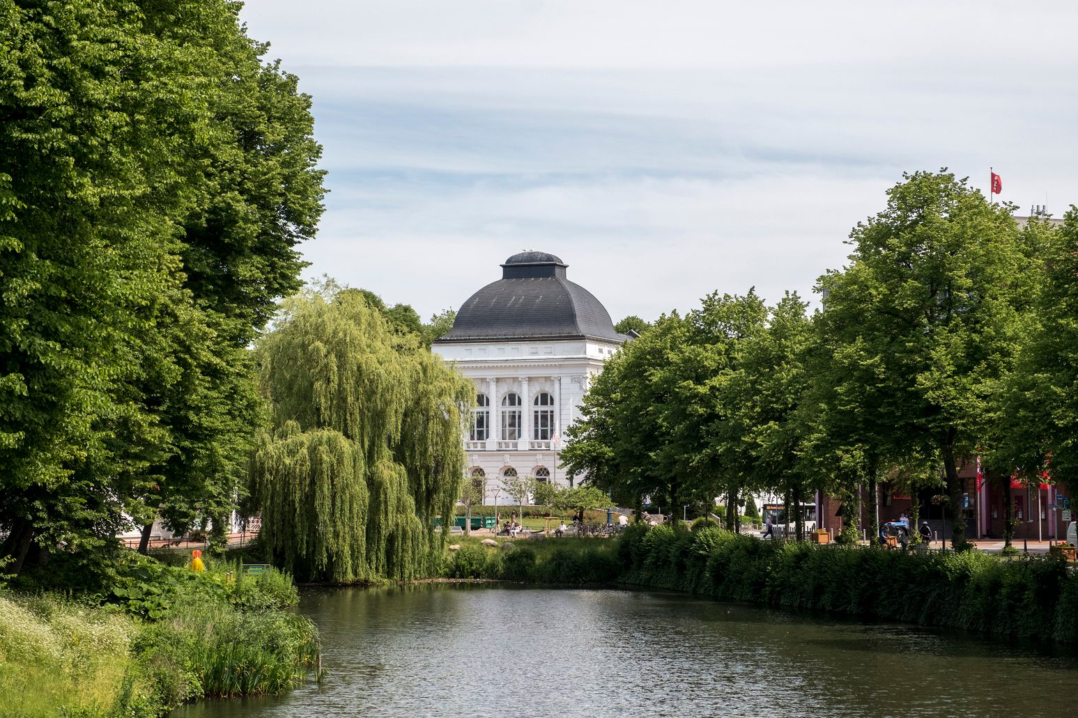 Das Stadttheater Rendsburg mit seiner markanten Kuppel, umgeben von üppigem Grün und einem Fluss im Vordergrund.