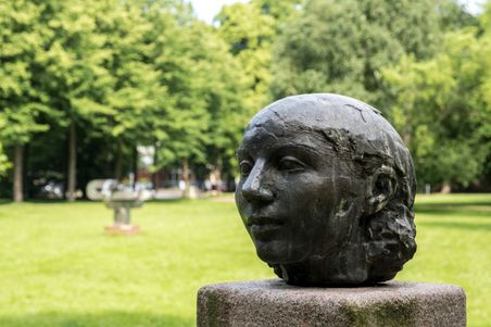 Bronzeskulptur eines Kopfes auf einem Sockel im Stadtpark Rendsburg, umgeben von grünen Bäumen und Rasenflächen.