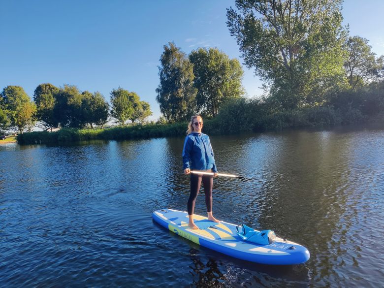 Eine Person steht auf einem Stand-Up-Paddleboard auf dem ruhigen Louisenbad. Im Hintergrund sind grüne Bäume und ein klarer blauer Himmel zu sehen.