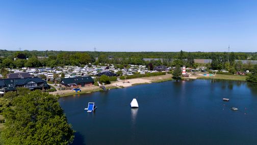 Luftaufnahme des Familien-Campingplatzes Padenstedt mit Wohnwagen, einem See, Strand und Spielplatz. Ein Leuchtturm und Wasserspielgeräte sind sichtbar.