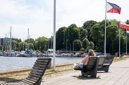 Eine Person entspannt auf einer Bank am Obereiderhafen in Rendsburg. Im Hintergrund sind Segelboote und mehrere Flaggen zu sehen.